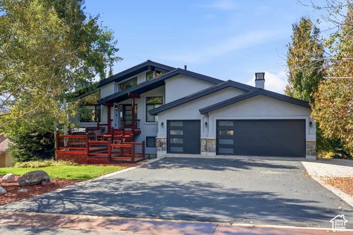 Contemporary exterior featuring multi-level rooflines, grey stucco siding, stone veneer accents, an elevated wooden deck, and two-car garage with frosted glass panel doors