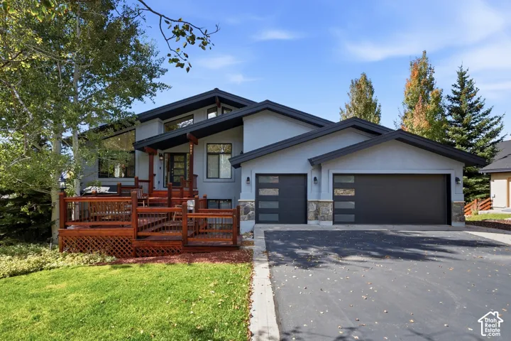 Split-level exterior featuring a multi-paneled wood deck, dual modern garage doors with horizontal glass insets, stone masonry accents, and expansive glass gables