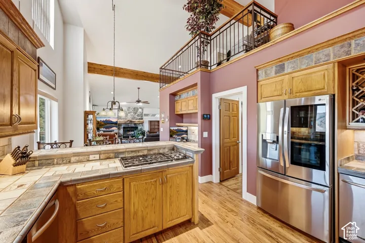 Kitchen featuring wood cabinetry, tile countertops with a built-in cooktop, stainless steel appliances, exposed wood beams, and a metal-railed mezzanine