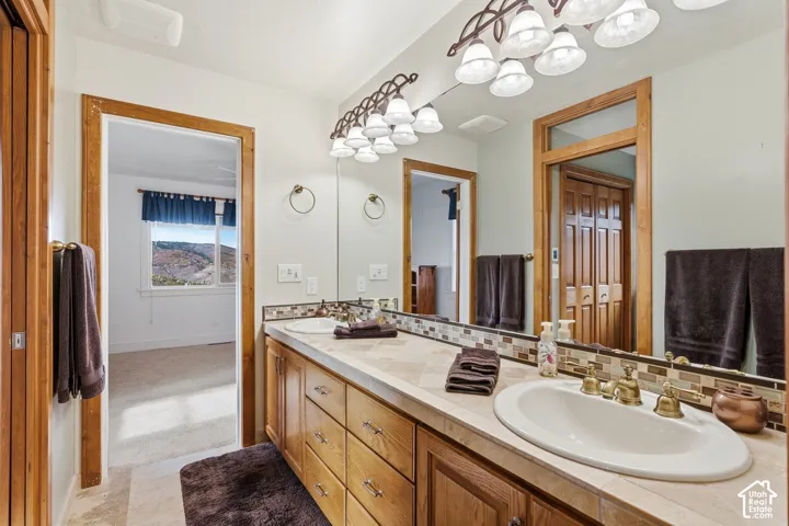 Dual-sink vanity with tile countertop, custom wood cabinetry, tile backsplash, and decorative brass-tone lighting fixtures