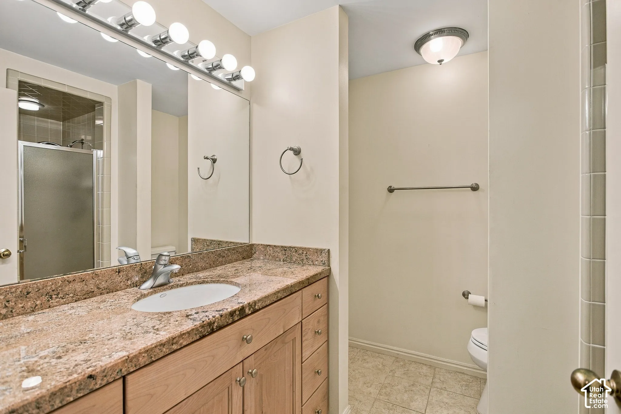 Bathroom featuring a wood vanity with stone-finish countertop, undermount sink, multi-bulb overhead lighting, and full-width mirror