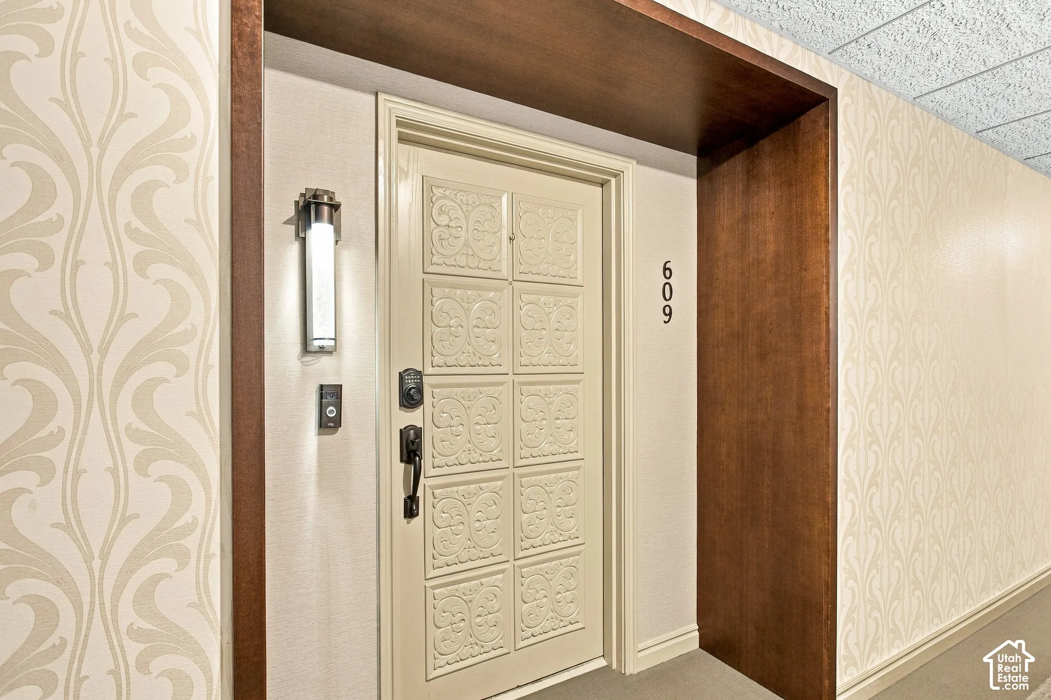 Entryway featuring a relief-patterned door, dark wood-finish door surround, integrated wall-mounted lighting, and patterned wallpaper