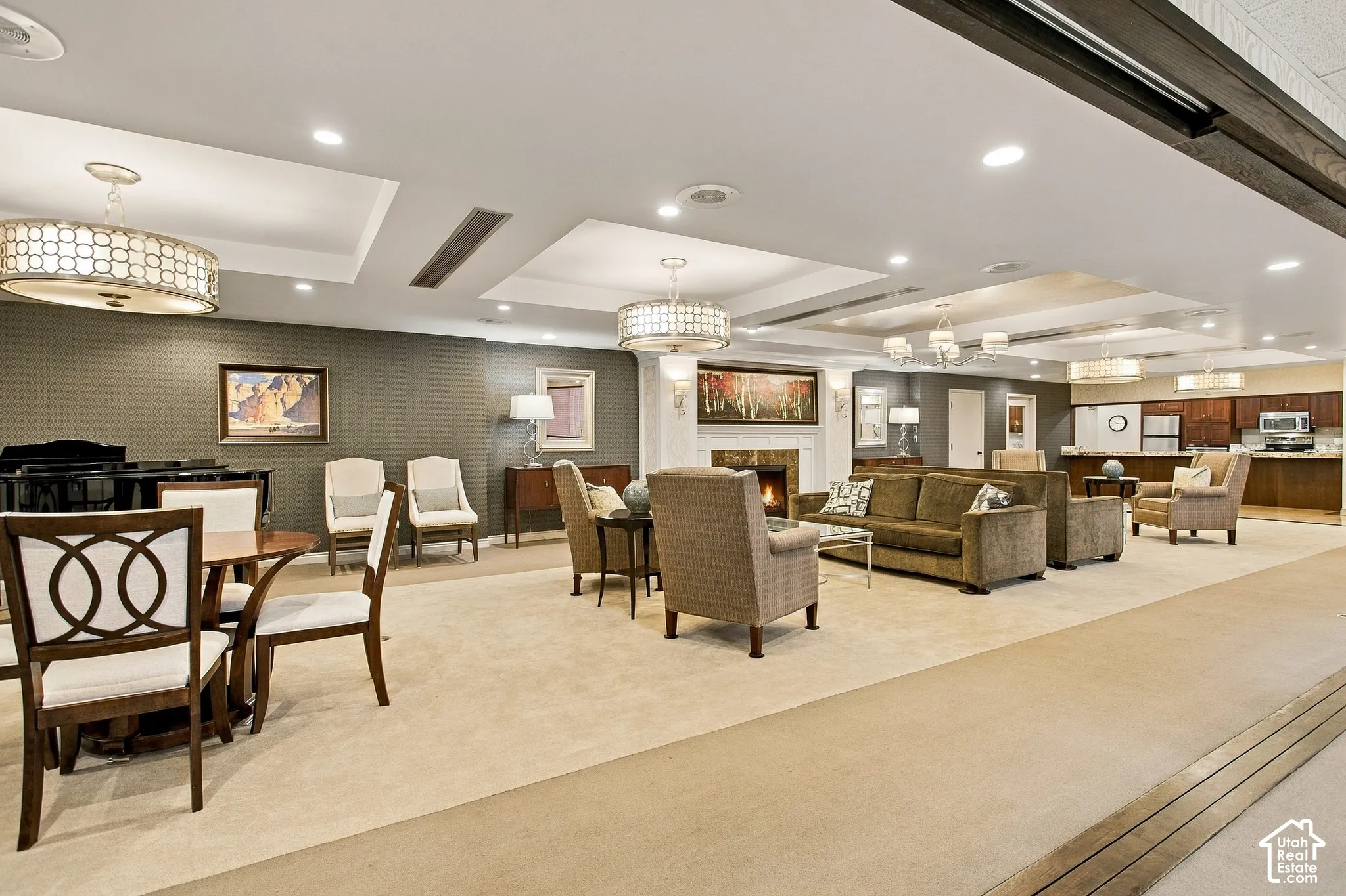 Expansive interior featuring recessed tray ceilings, multiple drum-style chandeliers, a built-in stone-surround fireplace, and a kitchenette with wooden cabinetry