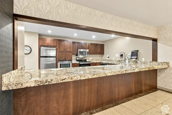 Pass-through counter with natural stone surfacing, wood-finish cabinetry, stainless steel appliances, recessed ceiling lighting, and patterned wall covering