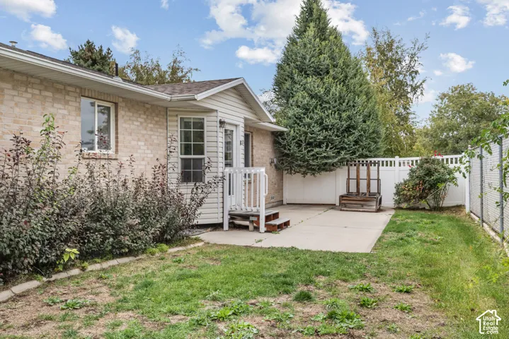 Exterior backyard featuring a concrete patio, light-toned masonry siding, white vinyl perimeter fencing, and established evergreen landscaping