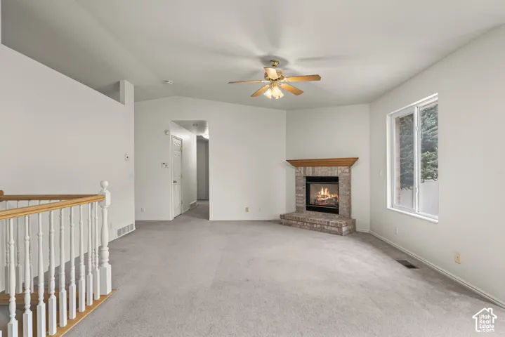 Vaulted ceiling, brick fireplace with a wood mantel, carpeted flooring, multi-light ceiling fan, and a staircase with a wooden handrail