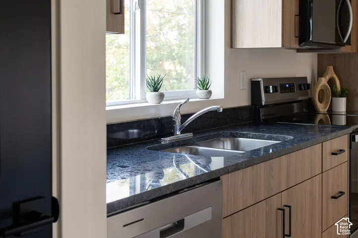 Kitchen featuring wood-finish cabinetry, dark stone-finish countertops, stainless steel undermount dual-basin sink, gooseneck faucet, and integrated stainless steel appliances