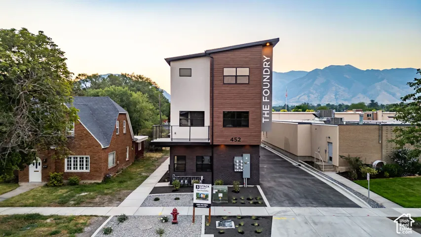 Modern multi-story exterior featuring a wood-finish siding, dark brick lower level, a mono-pitch roofline, and an integrated balcony with glass railing, set against a mountain backdrop