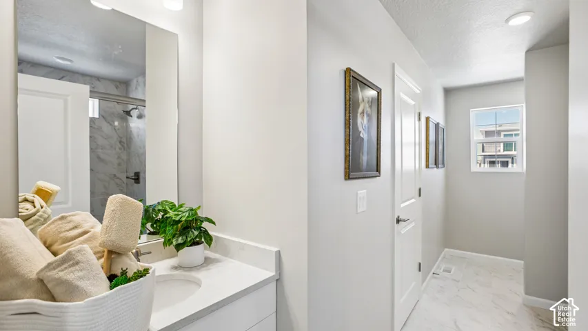 Bathroom featuring a vanity with a light-toned countertop, integrated basin, and full-height vanity mirror, stone-finish flooring, and a walk-in shower with light-toned wall tile