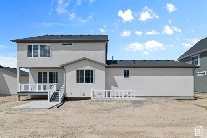 Exterior rear elevation featuring light-colored stucco siding, a grey shingle roof, a raised wooden deck with white railing, and a concrete patio