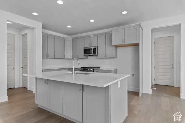 Kitchen featuring grey shaker-style cabinetry, quartz countertops, a central island with a stainless steel undermount sink, wood-finish flooring, and recessed ceiling lighting