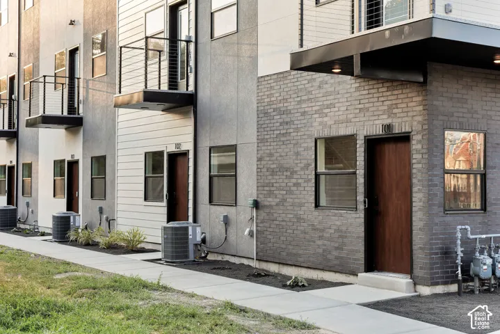 Modern multi-story exterior featuring a gray brick facade, horizontal siding, individual elevated metal balconies, and dark-finished doors