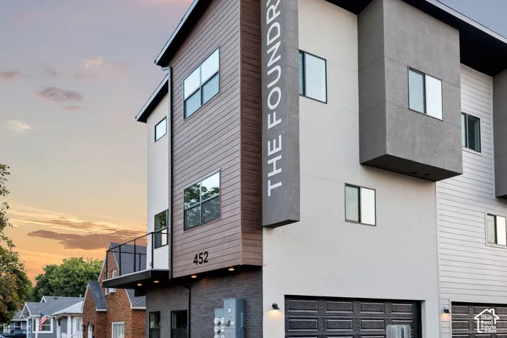 Modern exterior facade featuring mixed materials of wood-finish siding, smooth stucco, and brick masonry, accompanied by a metal-railed balcony and multi-level geometric design