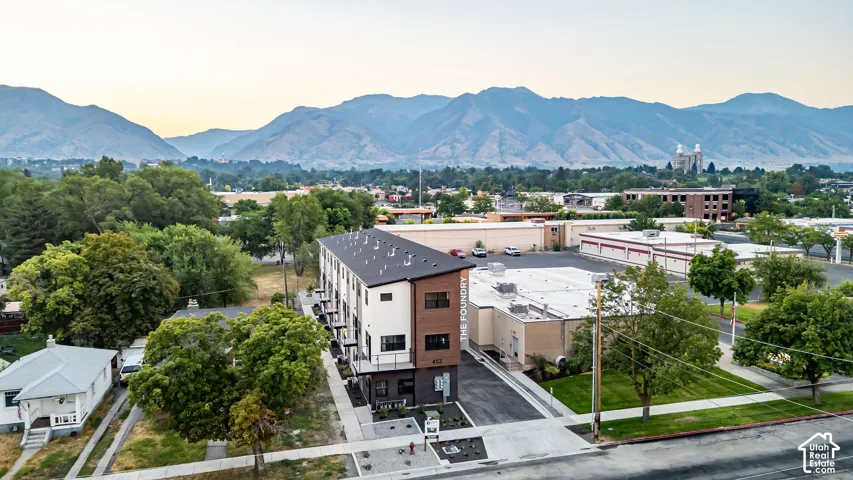 Multi-story building with a shed roof, light-toned stucco and wood-finish siding, private balconies, and asphalt-paved drive, set against a backdrop of mountains
