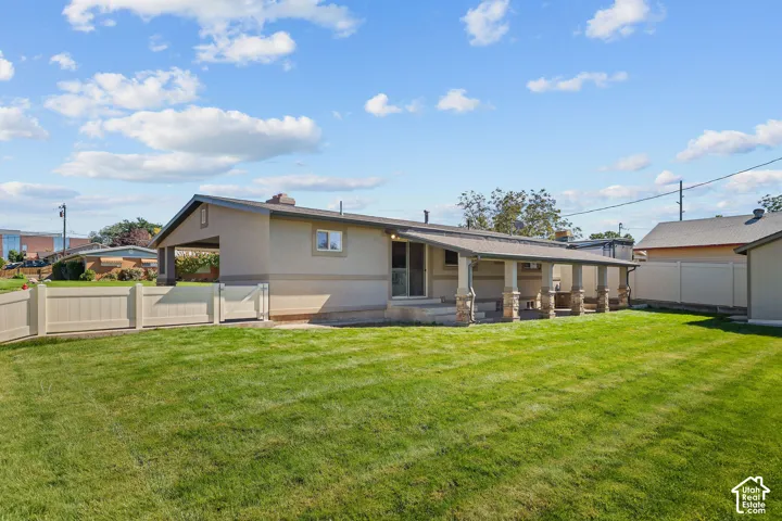 Rear exterior featuring a covered patio with stone-accented support columns, expansive lawn, vinyl fencing, and a side carport