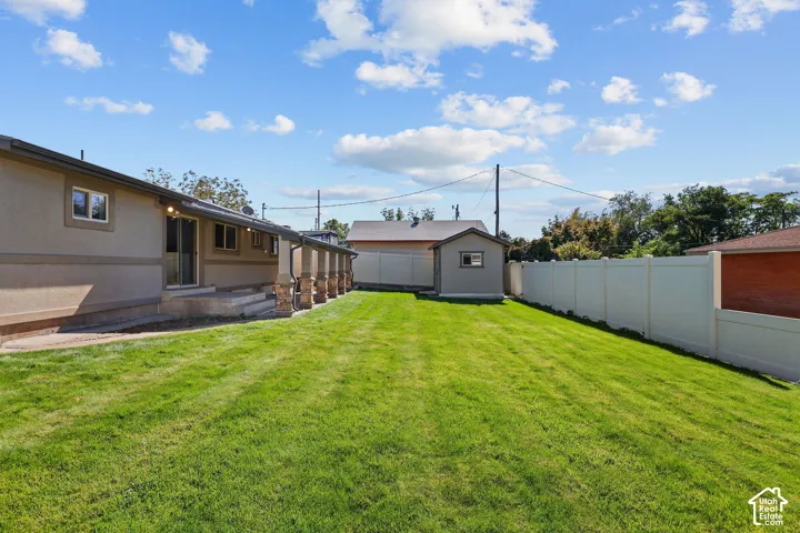 Backyard featuring a manicured lawn, covered concrete patio with stone-accented columns, detached storage structure, and perimeter vinyl privacy fencing