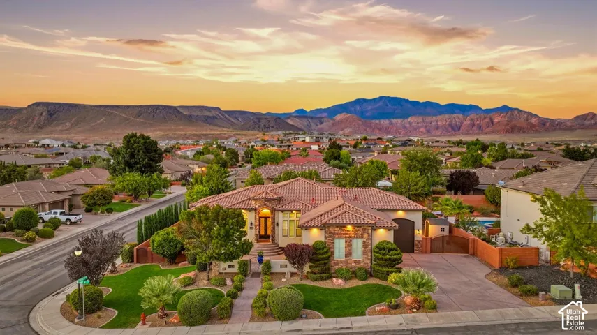 Single-level residence featuring a clay tile roof, stone masonry facade, arched entryway, paved driveway, and manicured landscaping set against a mountain backdrop