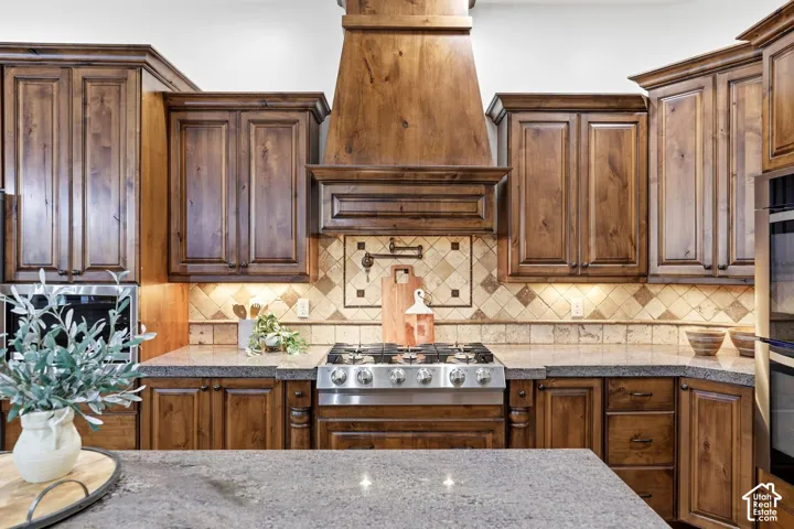 Custom wood cabinetry, stainless steel gas range, stone-tiled backsplash with a wall-mounted pot filler, professional-grade range hood, and stone countertops