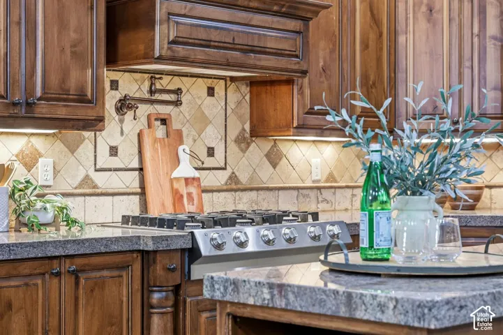 Custom stained wood cabinetry, stone countertops, professional-grade gas range, tiled backsplash with decorative inset, and wall-mounted pot filler