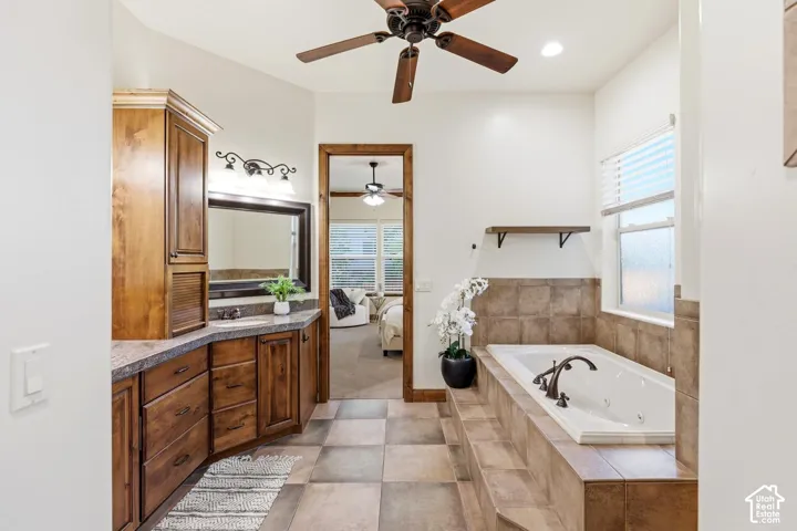 Custom wood cabinetry, jetted soaking tub with tile surround, large-format tile flooring, bronze-finish fixtures, and ceiling fan