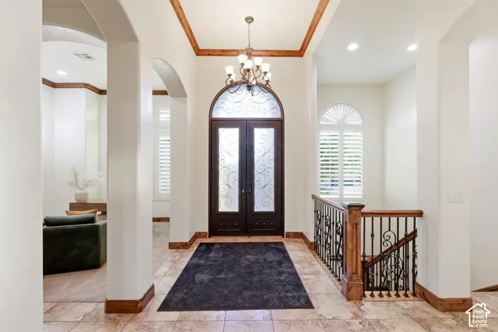 Foyer featuring arched double doors with decorative metal inlays, tile flooring, crown molding, iron-detail railing, and a multi-arm chandelier