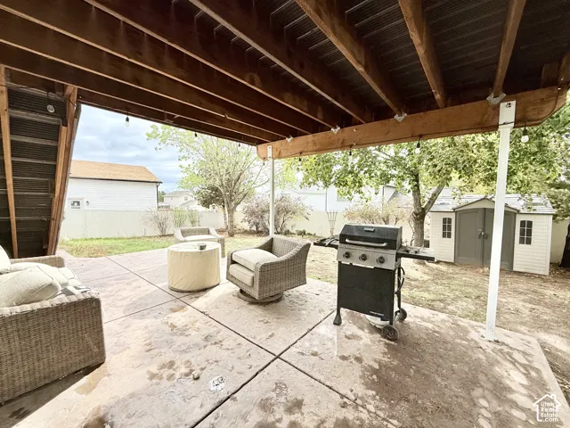 Covered concrete patio featuring exposed wood beam framing, recessed string lighting, and an enclosed storage shed within the backyard