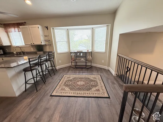 Kitchen area featuring wood-finish flooring, a stone-surfaced island with bar seating, white cabinetry, a bay window with integrated horizontal blinds, and a metal-railed staircase