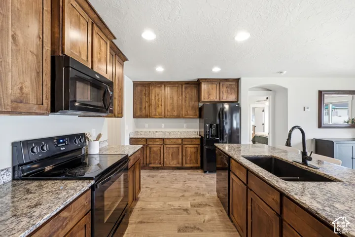 Kitchen featuring stained wood cabinetry, stone-pattern countertops, integrated black appliances, a central island with an undermount sink, and wood-finish flooring