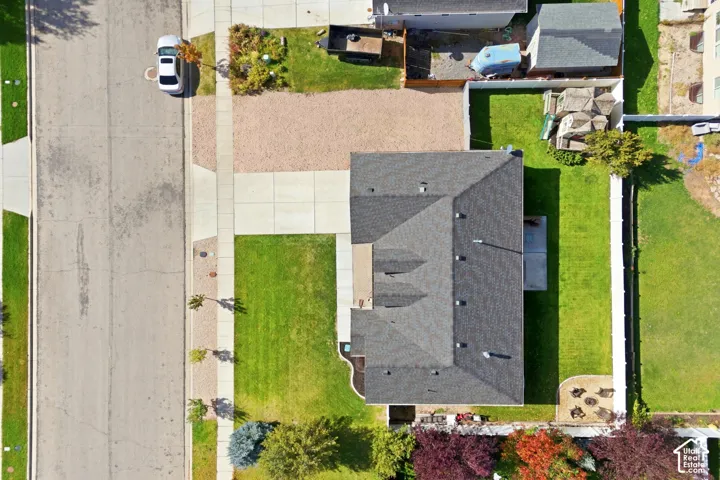 Aerial perspective of a property featuring a concrete driveway, gravel parking area, shingle roof, enclosed lawn, and perimeter fencing