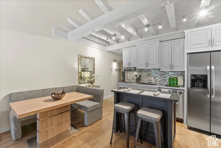 Kitchen featuring white shaker-style cabinetry, a dark-toned center island with an integrated breakfast bar, stone-tile backsplash, stainless steel refrigerator, and exposed ceiling beams