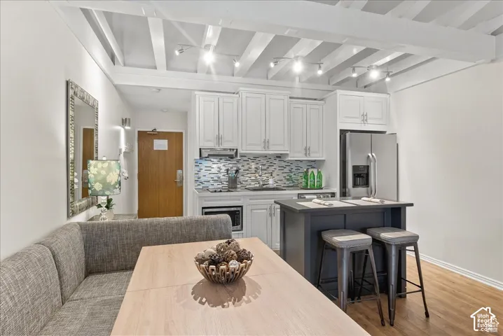 Kitchen featuring white cabinetry, a dark-finish central island, mosaic tile backsplash, wood-finish flooring, and exposed ceiling beams