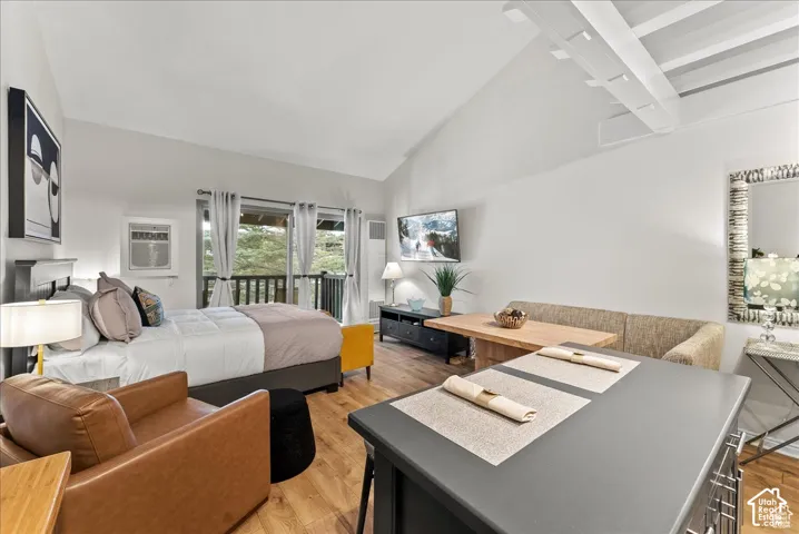 Vaulted ceilings with exposed white structural beams, wood-finish flooring, and sliding glass doors with access to a railed balcony