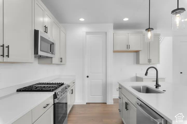 Kitchen featuring light gray shaker-style cabinetry, quartz countertops, stainless steel appliances, a center island with an undermount sink and matte black hardware, and wood-finish flooring