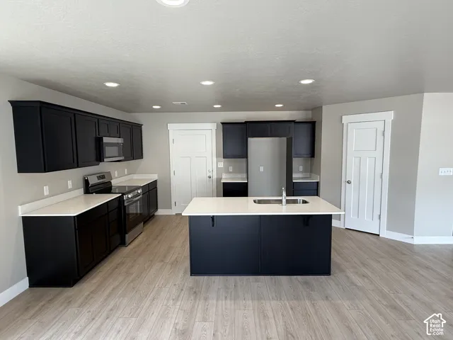 Kitchen featuring dark cabinetry, solid-surface countertops, center island with integrated sink, stainless steel appliances, and wood-finish flooring