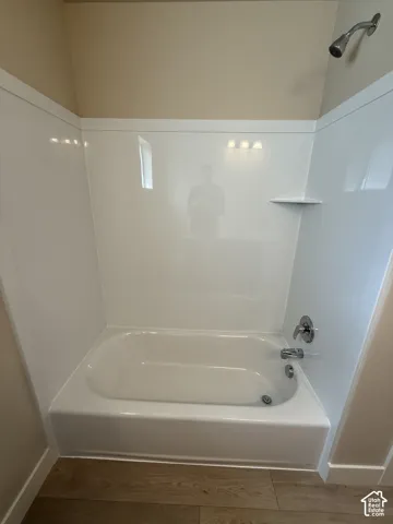 Bathtub featuring a white molded surround, chrome shower head, integrated shelving, and polished chrome faucet hardware, finished with wood-finish flooring