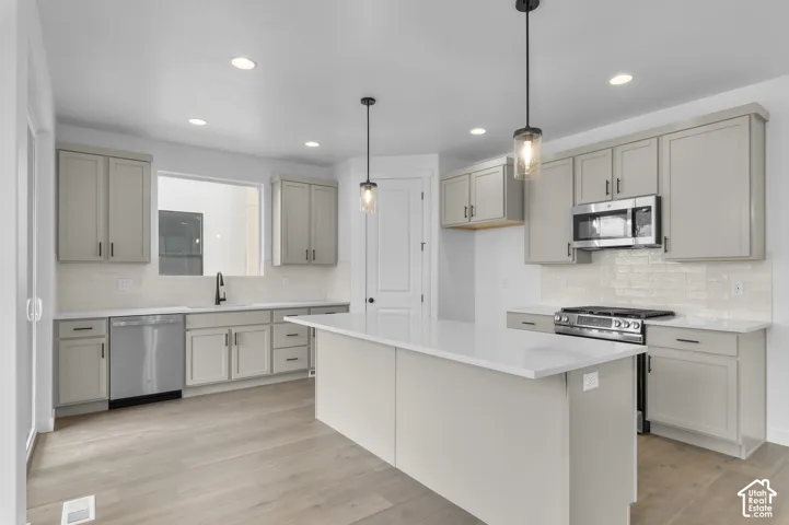 Kitchen featuring light gray cabinetry, a white countertop island, stainless steel appliances, a light-toned tile backsplash, and wood-finish flooring