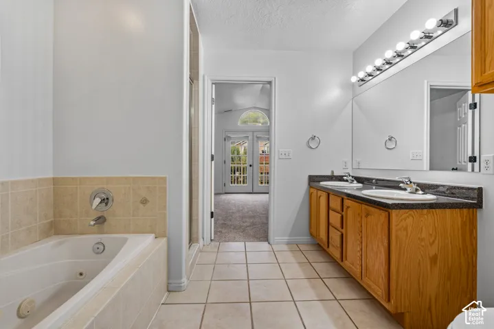 Dual-sink vanity with dark stone countertop, large wall-mounted mirror, soaking tub with tile surround, and ceramic tile flooring