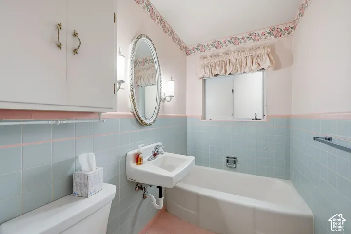 Bathroom featuring pastel-toned ceramic wall tiling, a wall-mounted sink, an oval wall mirror with decorative frame, built-in upper cabinetry, and a sliding window with a valance