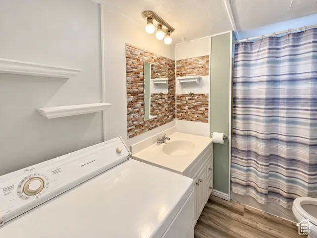 Bathroom featuring wood-finish flooring, a vanity with a solid-surface countertop and integrated sink, decorative stacked-stone backsplash, wall-mounted shelving, and a three-bulb vanity light fixture