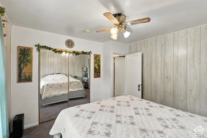 Neutral-toned wood-finish paneling, mirrored sliding closet doors, carpeted flooring, and a ceiling-mounted light fixture with a multi-blade fan