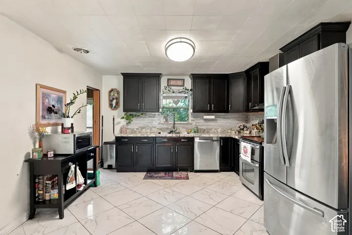 Kitchen featuring dark-finish cabinetry, stone-style countertops, neutral tile flooring in a diamond pattern, stainless steel appliances, and a horizontal-tiled backsplash
