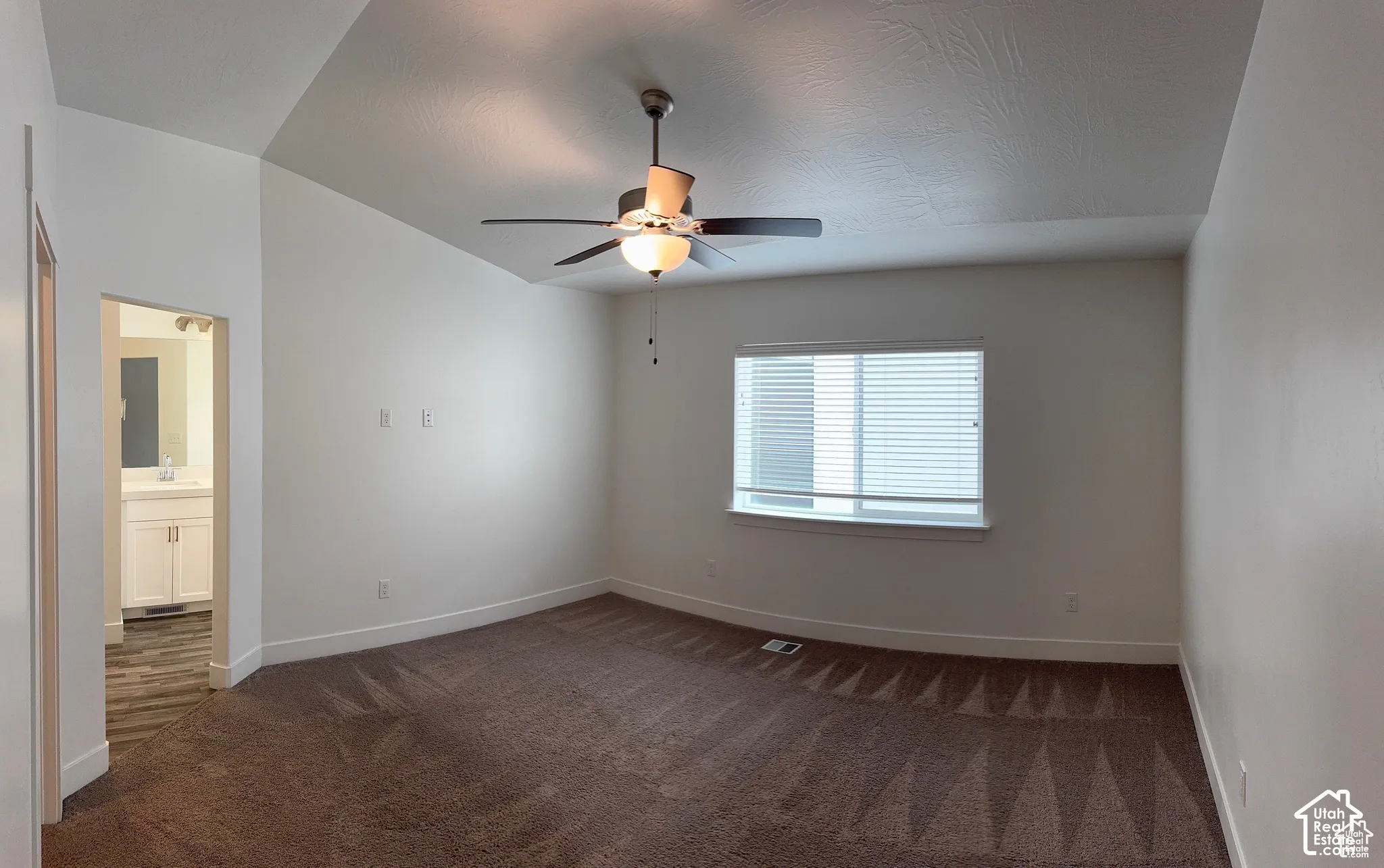 Vaulted ceiling, ceiling fan with integrated lighting, window with blinds, brown carpet flooring, and an adjoining bathroom featuring a white vanity with an undermount sink