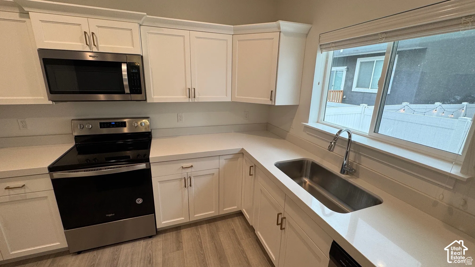 White shaker-style cabinetry with gold-tone hardware, stainless steel appliances, a large stainless steel sink with a gooseneck faucet, light-toned countertops, and wood-finish flooring