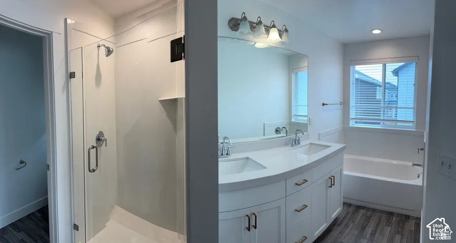 Dual vanity with integrated sinks, wood-finish flooring, a glass-enclosed shower, a built-in bathtub, and a window with horizontal blinds