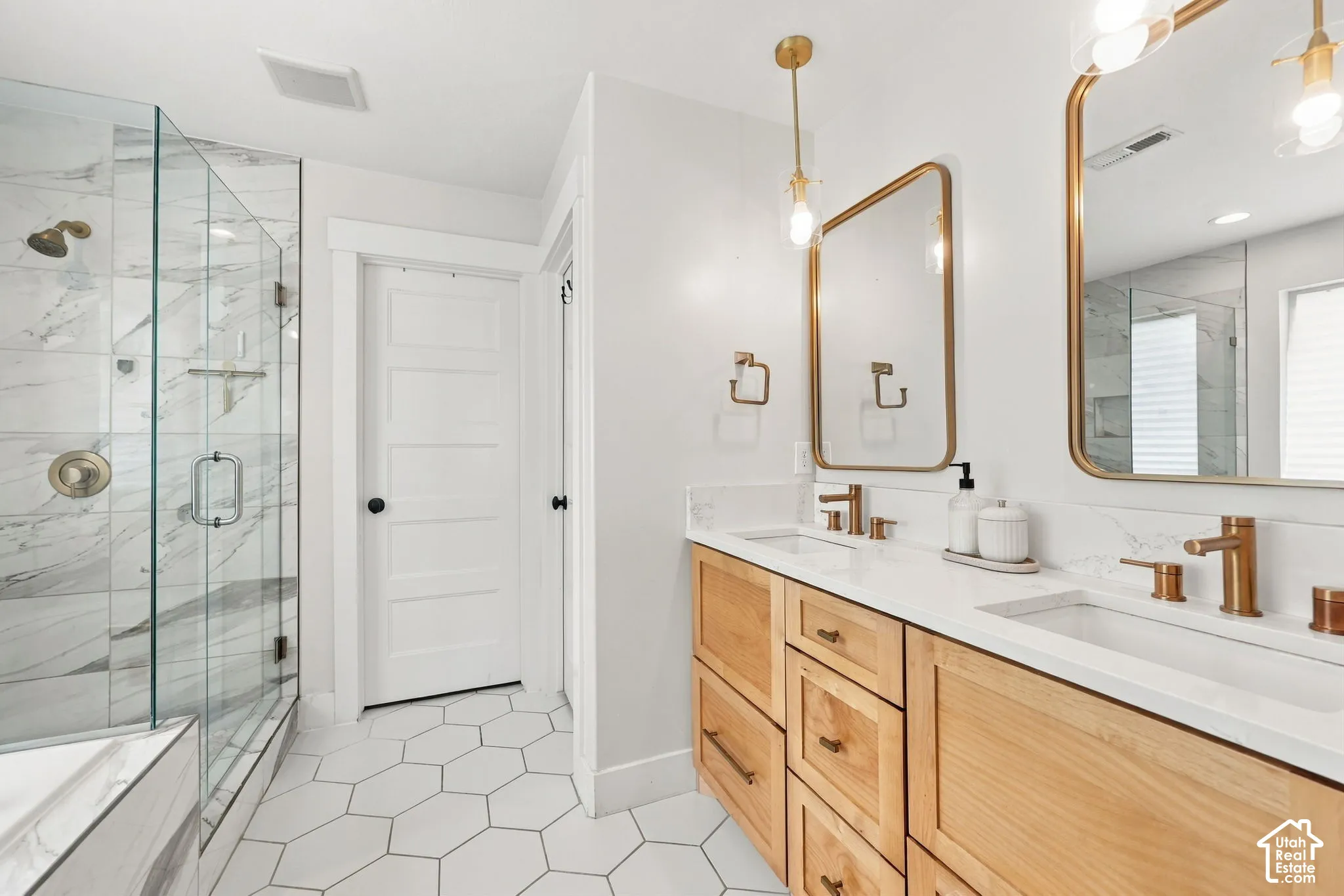 Bathroom featuring a dual-sink wood-finish vanity with a white countertop, hexagon tile flooring, glass-enclosed shower with tiled surround, and gold-toned fixtures