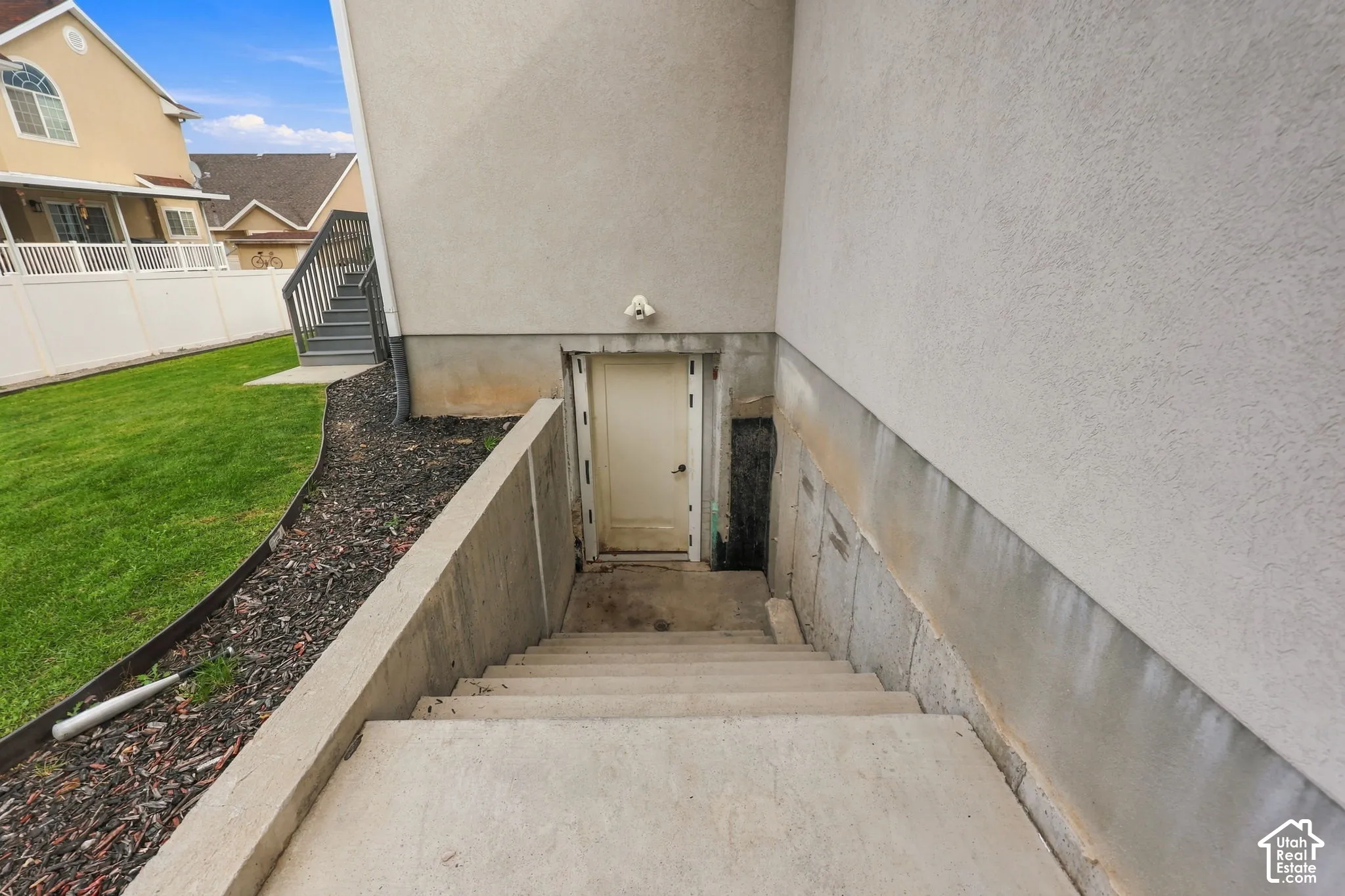 Exterior concrete staircase leading to a recessed door, stucco siding, and a landscaped yard with mulched garden beds and a white vinyl privacy fence