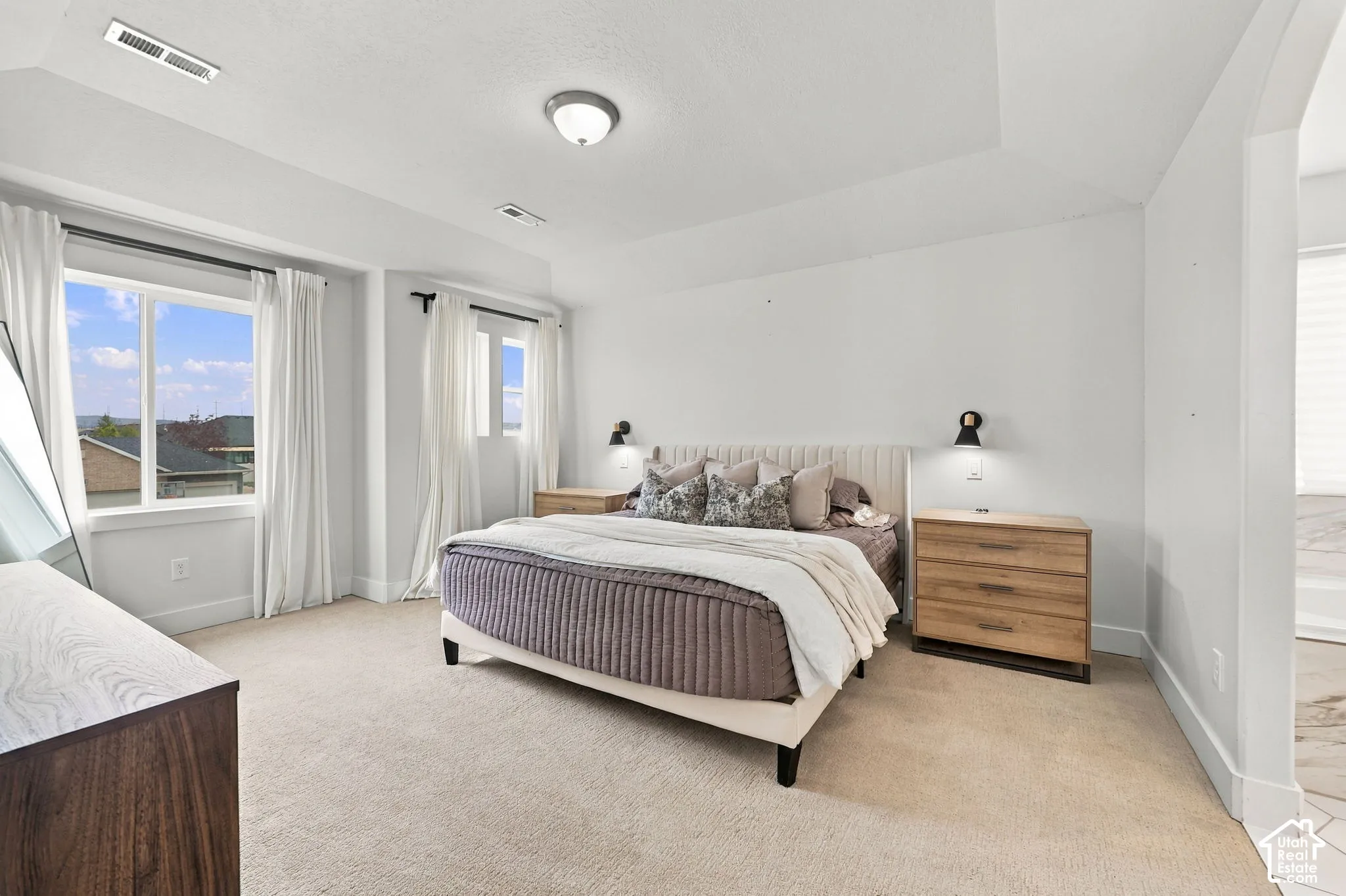 Tray ceiling with central flush-mount lighting, wall-mounted side sconces, multiple windows with neutral-toned trim, and wall-to-wall carpeting