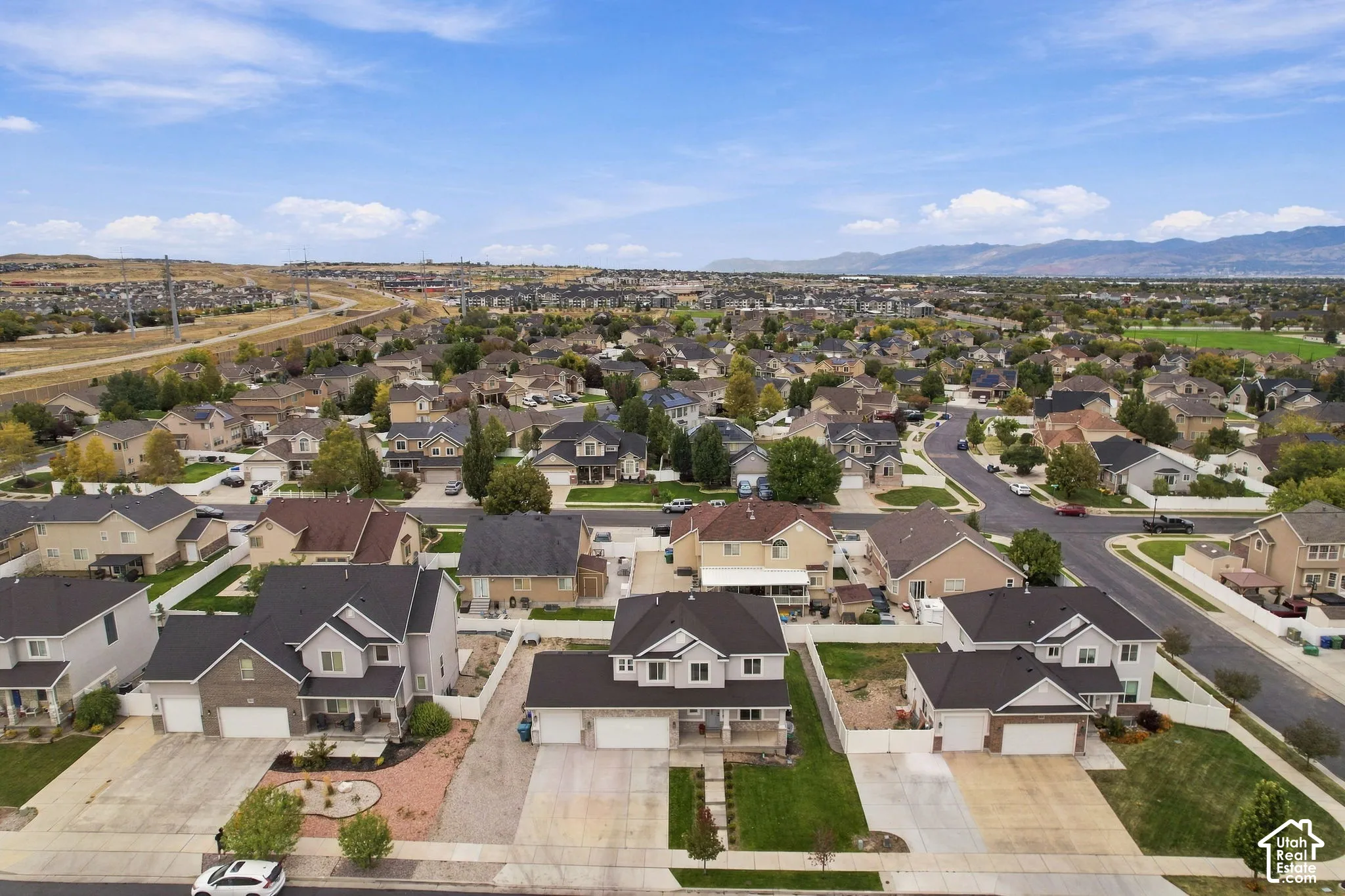Aerial perspective of a multi-story residential property featuring a three-car garage, concrete driveway, front lawn landscaping, and white perimeter fencing