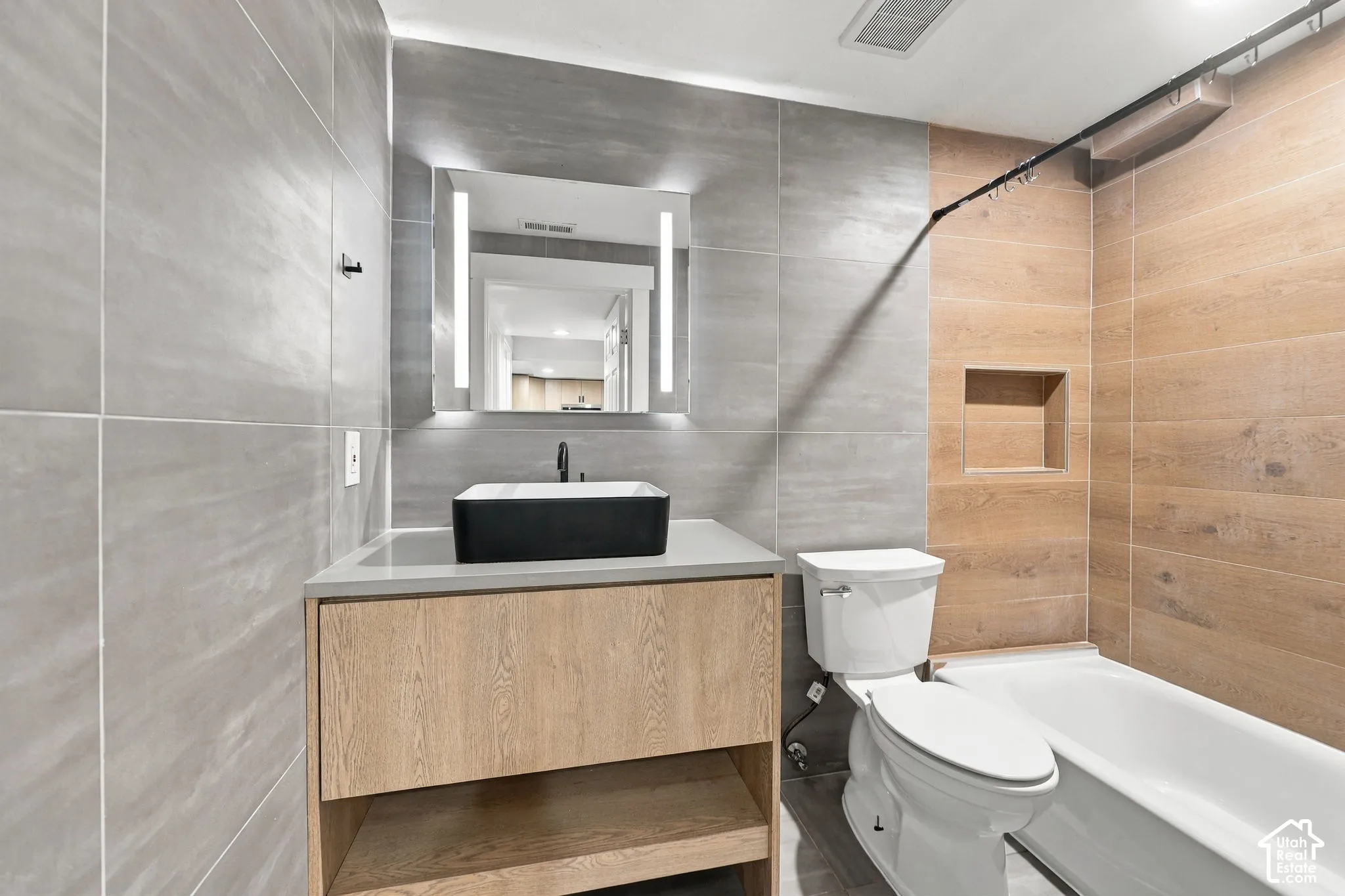 Bathroom featuring a floating wood-finish vanity, rectangular vessel sink, LED-integrated mirror, large-format grey tile walling, and wood-finish tile tub surround with a built-in recessed niche