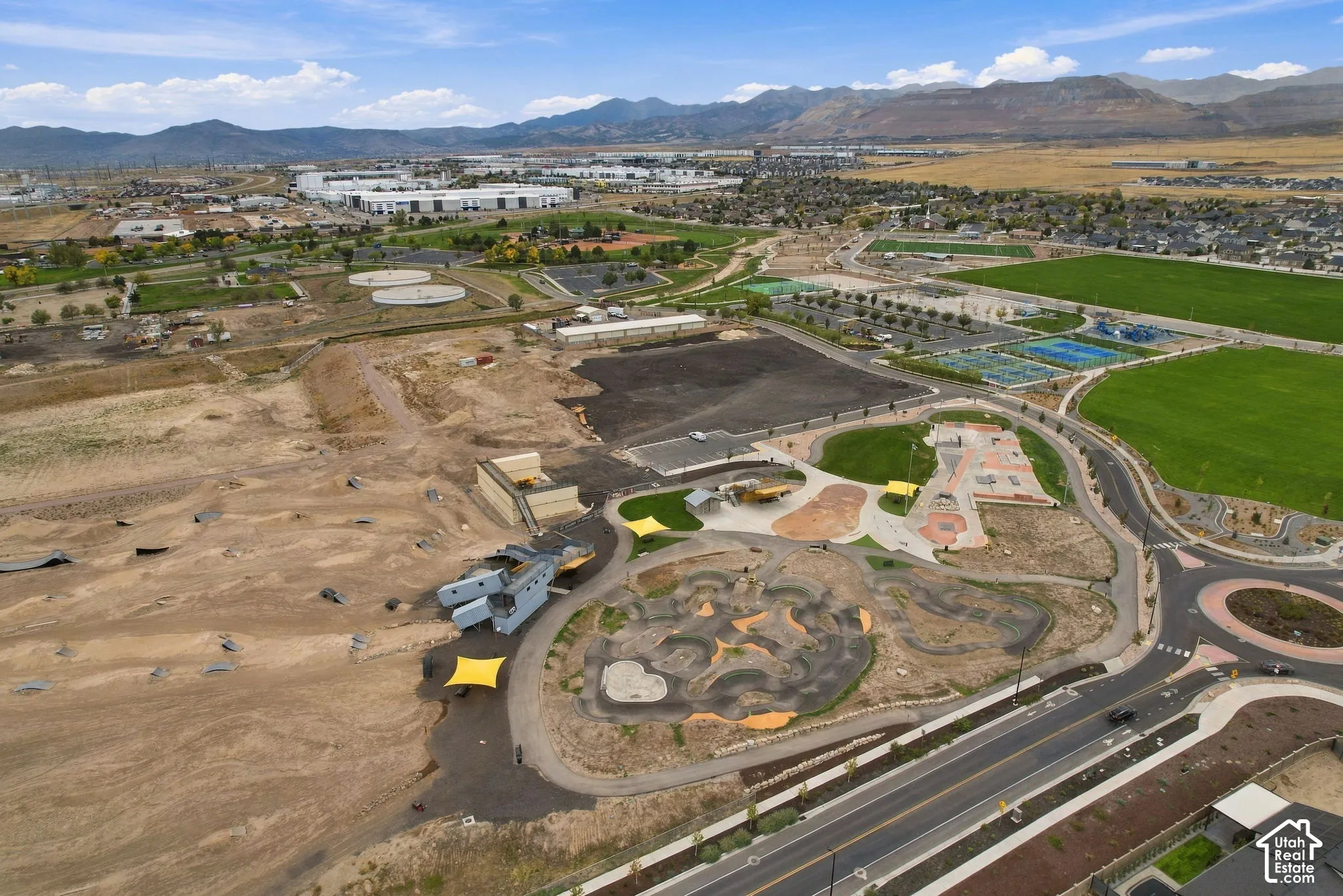 Aerial perspective of a park area featuring a paved pump track, concrete skate park, multi-use turf fields, and mountain backdrop